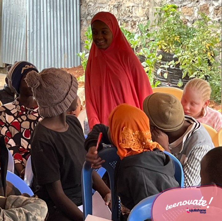 A young woman in a red hijab speaking to a seated group of girls