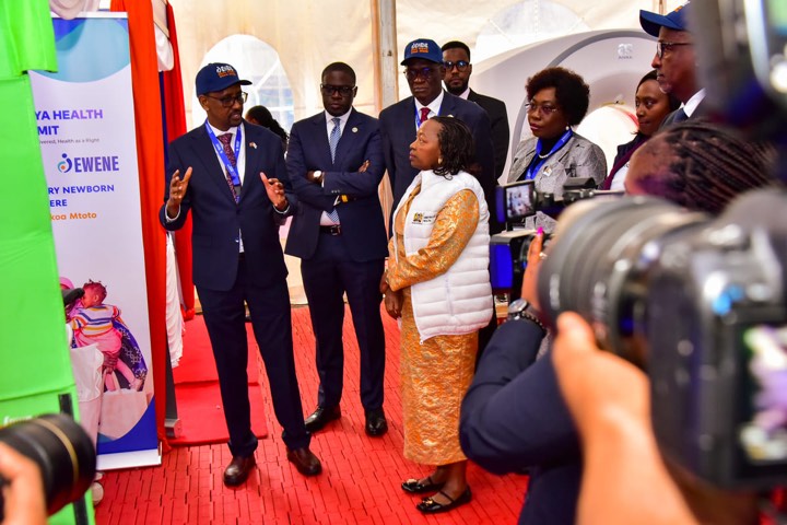 Officials in suits and EWENE caps touring an exhibition stand as photographers look on