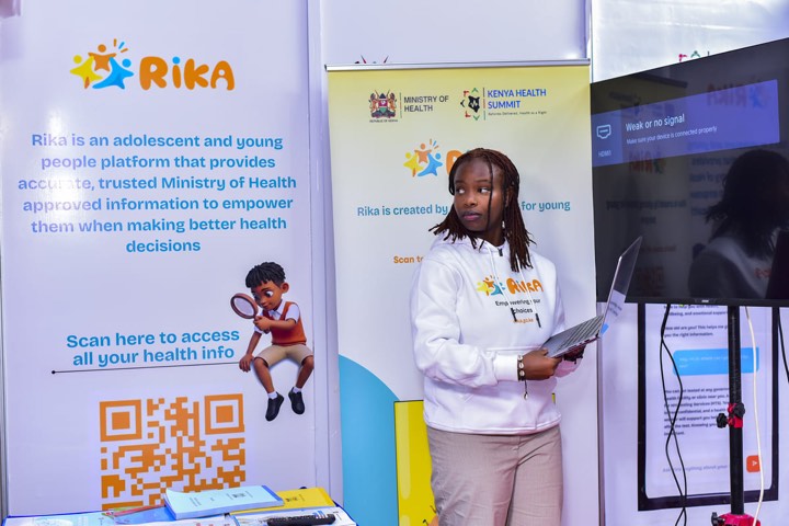 A young woman in a RIKA hoodie standing beside the RIKA banner