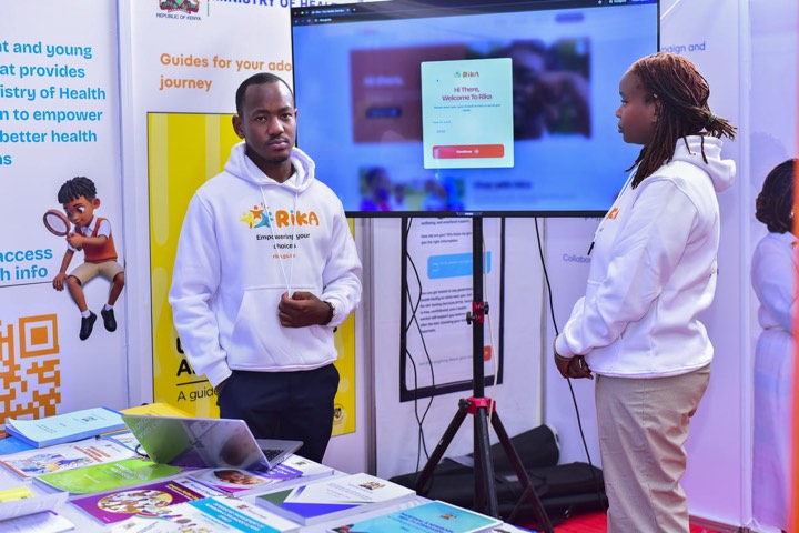 Two young people in RIKA hoodies beside a screen showing the RIKA platform