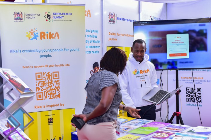 A young man in a RIKA hoodie showing a laptop to a visitor at the RIKA stand