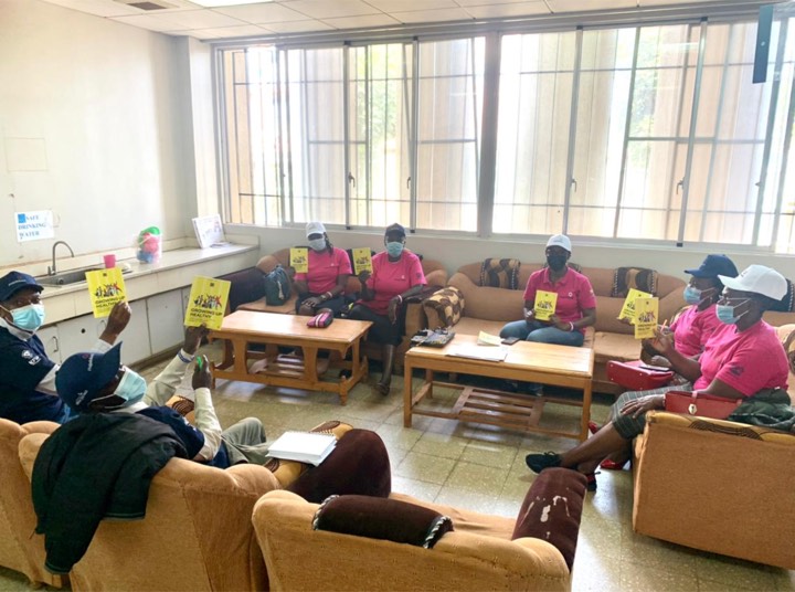 Women in pink Tujulishane shirts holding up yellow booklets during a meeting